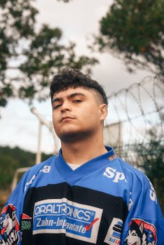 Confident young man in a blue sports jersey standing outside, exuding a cool and bold demeanor.