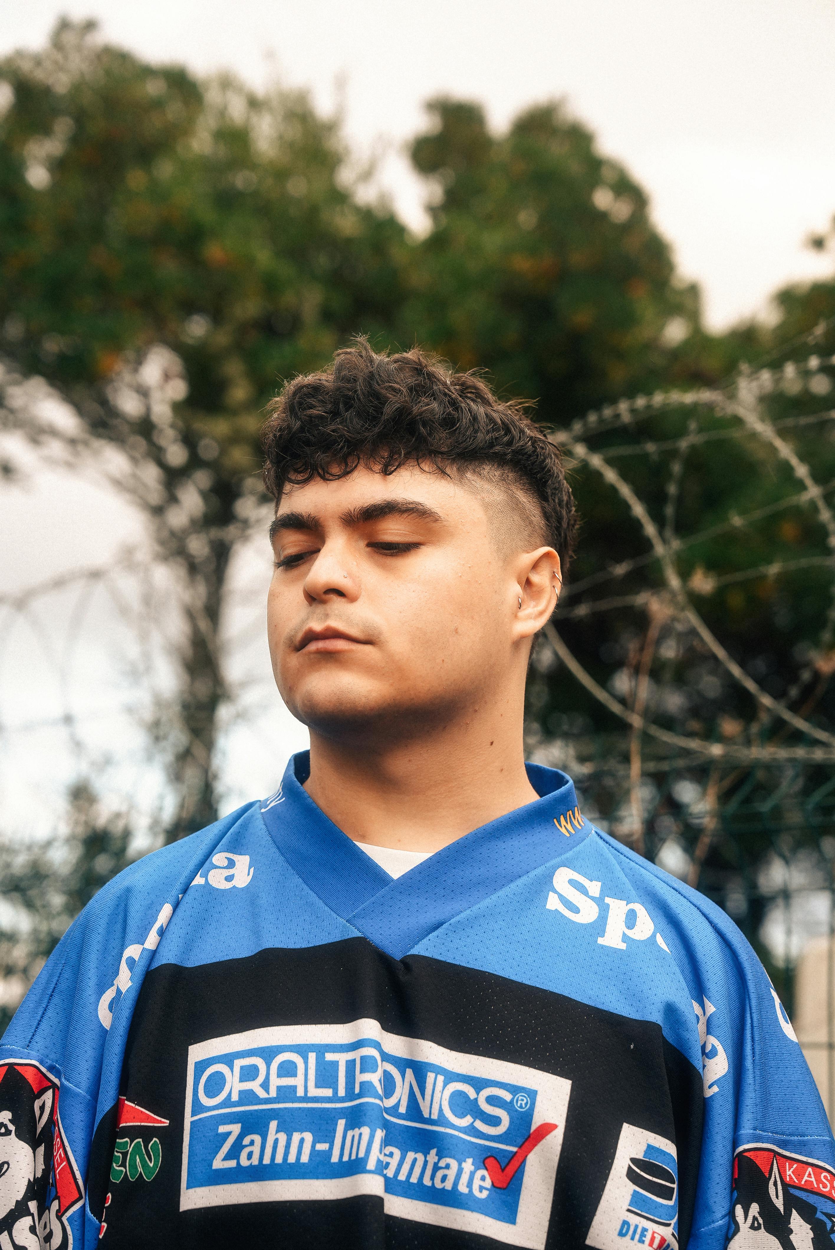 Portrait of young man in a vibrant blue jersey with eyes closed, standing outdoors against a barbed wire fence.