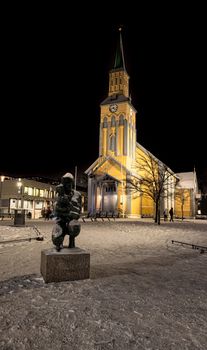 Tromsø Cathedral illuminated against a snowy winter night sky, featuring a nearby statue.
