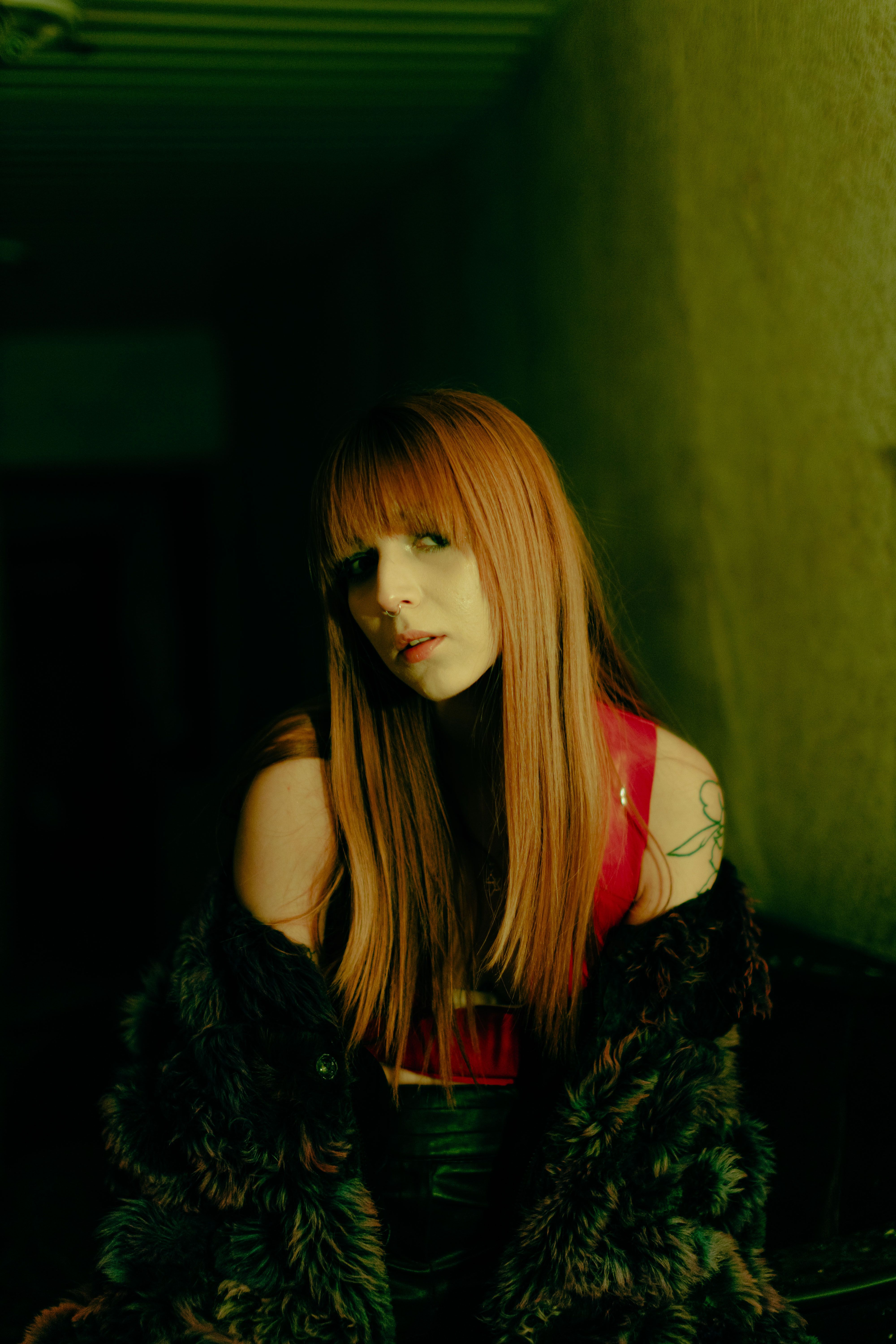 Portrait of a woman with red hair and tattoos in a dimly lit hallway, exuding a moody atmosphere.