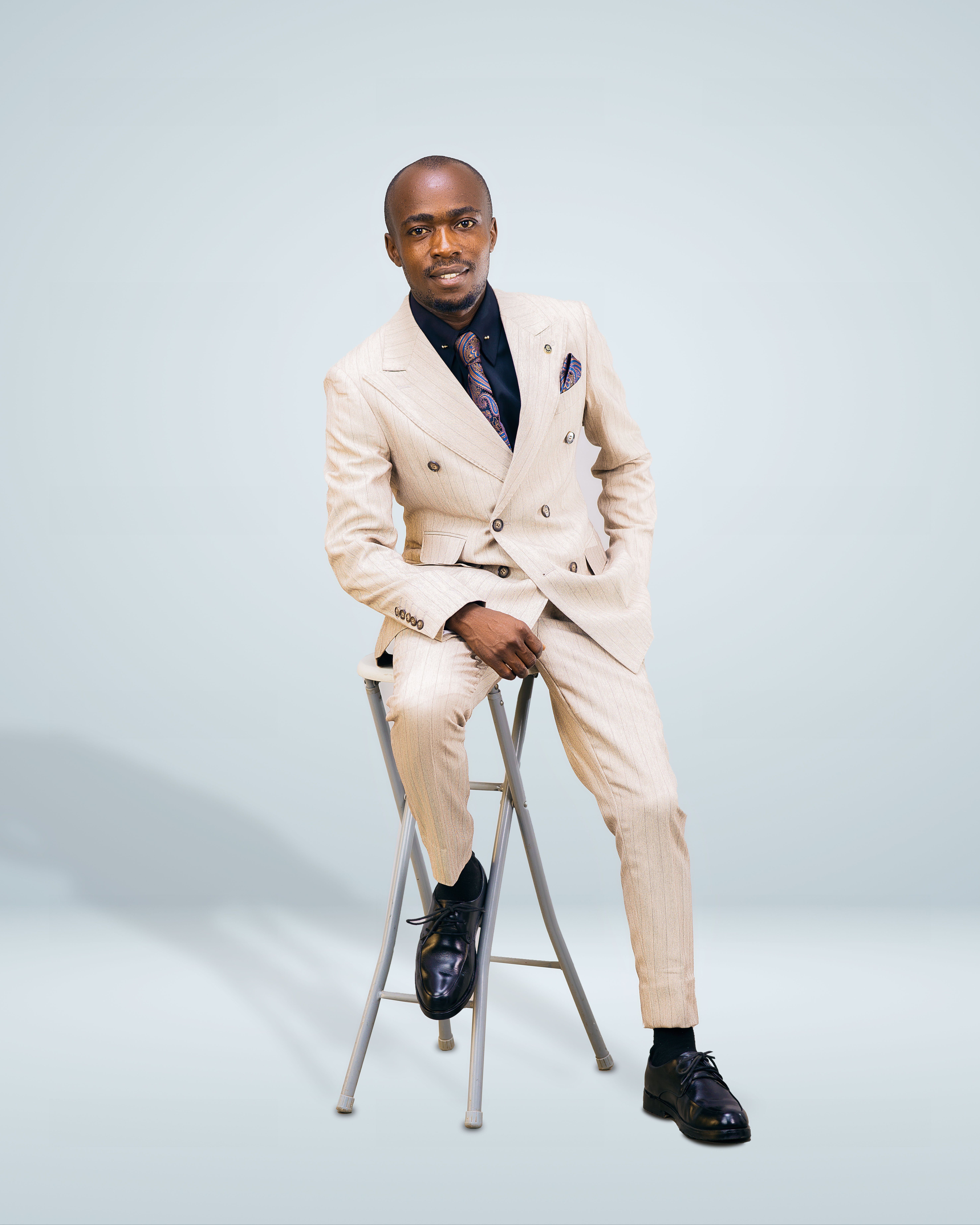 Confident man in beige suit sitting on a stool against a plain background.