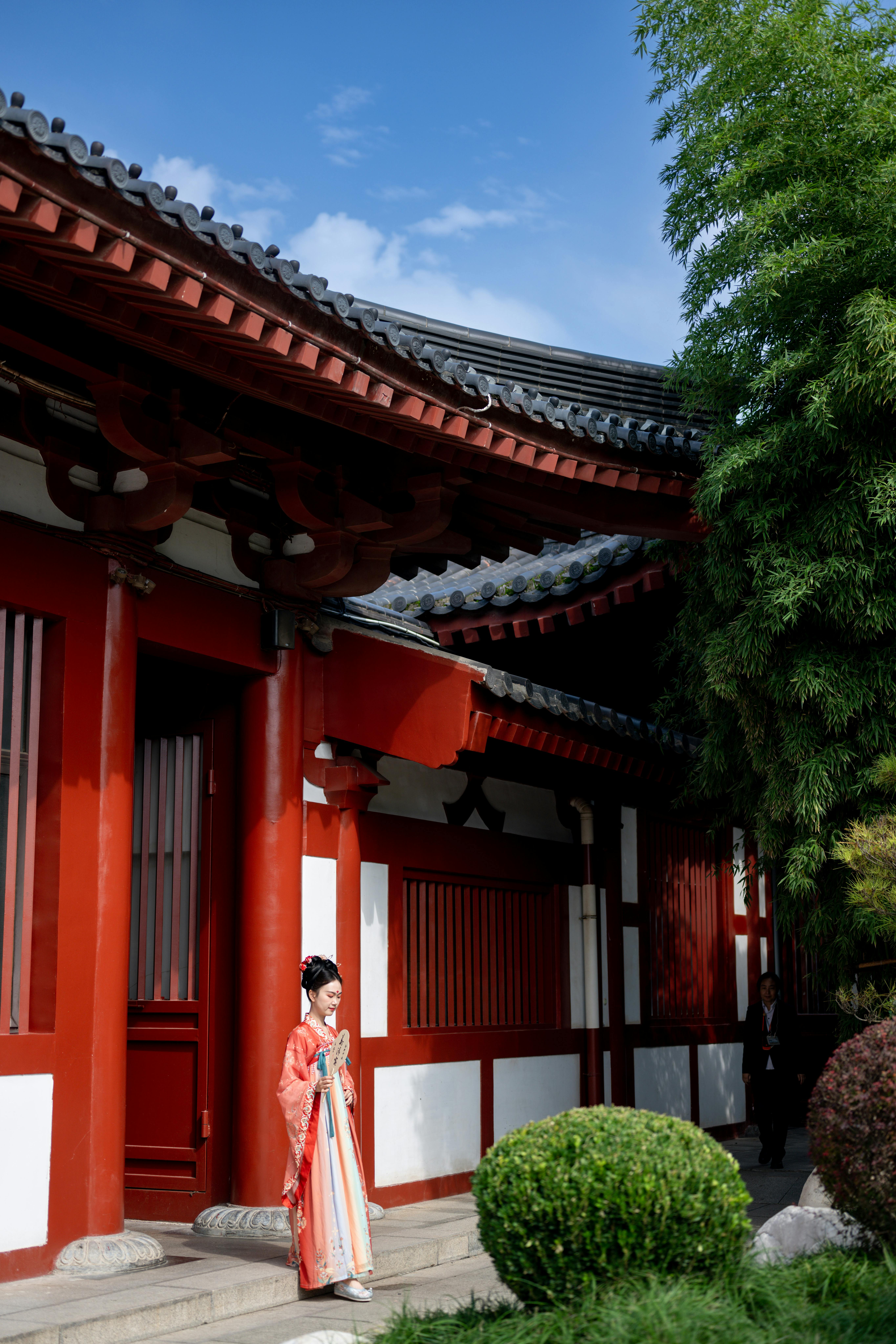A woman in a kimono stands by traditional Japanese architecture on a sunny day.