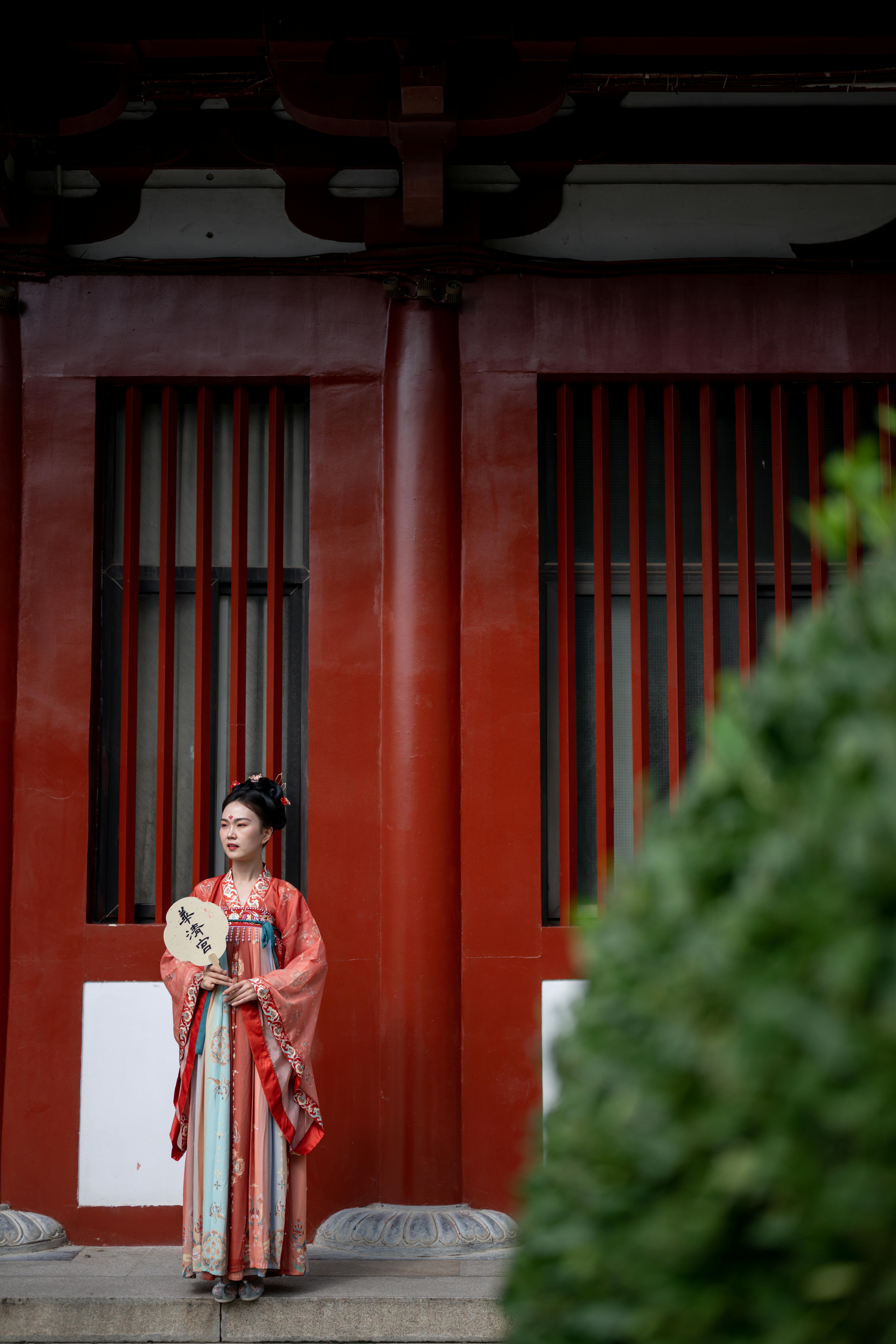 Woman in traditional hanfu dress holding a fan, standing by a red-pillared building.