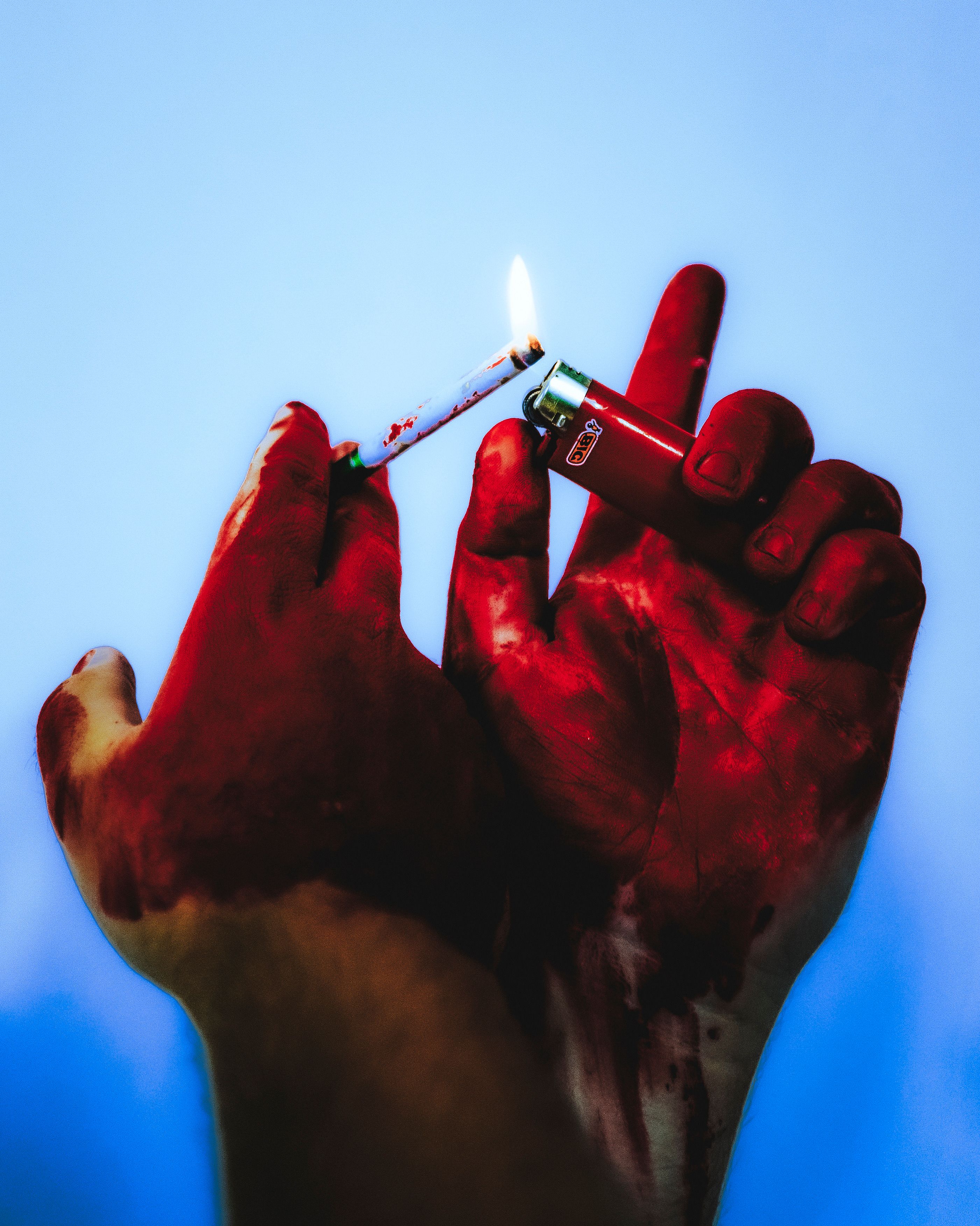 Close-up of red-stained hands lighting a cigarette with a red lighter against a blue background.