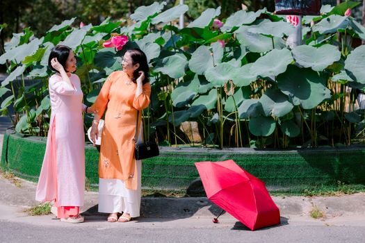 Two women in traditional Asian dresses converse beside a lush lotus pond with a red umbrella.