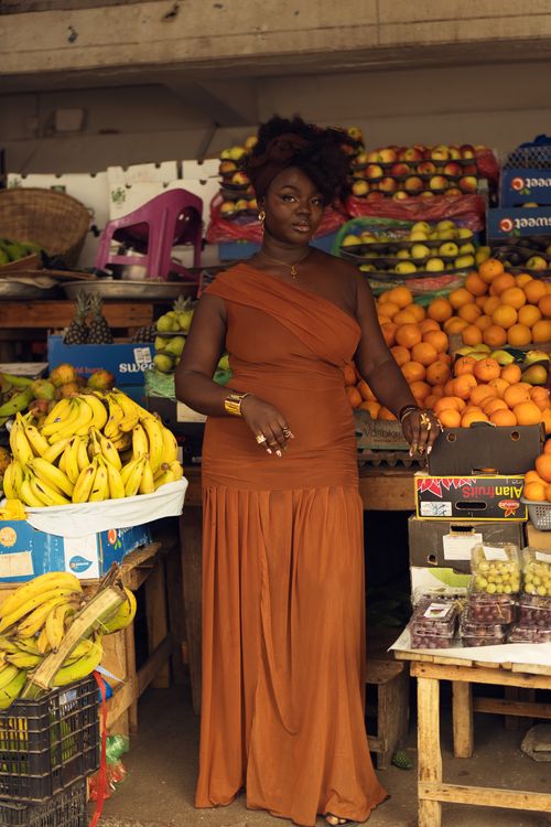 Free Elegant woman in orange dress poses amidst vibrant fruit market display. Stock Photo