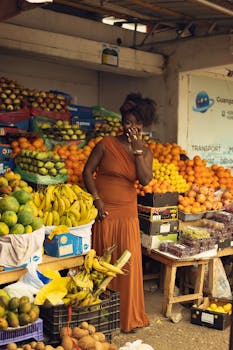 A woman in a rust-colored dress stands amidst vibrant fruits at a busy market.