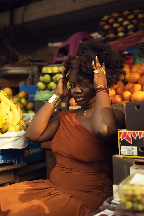 Free Stylish woman in orange dress poses amidst colorful fruit at an outdoor market. Stock Photo