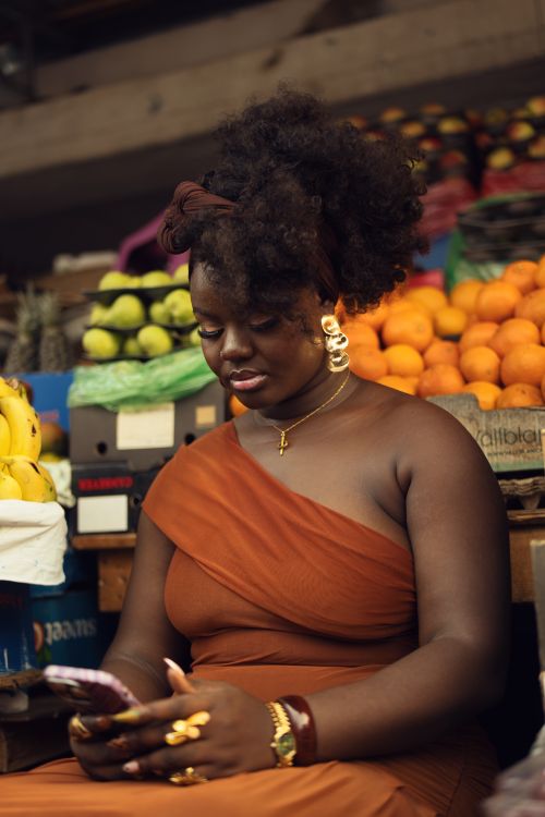 Free Woman in a vibrant orange dress uses smartphone in a market surrounded by colorful fruits. Stock Photo