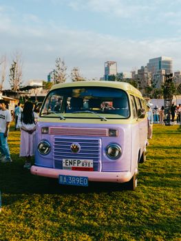 A colorful vintage Volkswagen van parked at a lively outdoor festival with people and cityscape in the background.