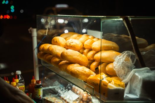 Evening street food cart displaying fresh baguettes and condiments, illuminated under warm light.