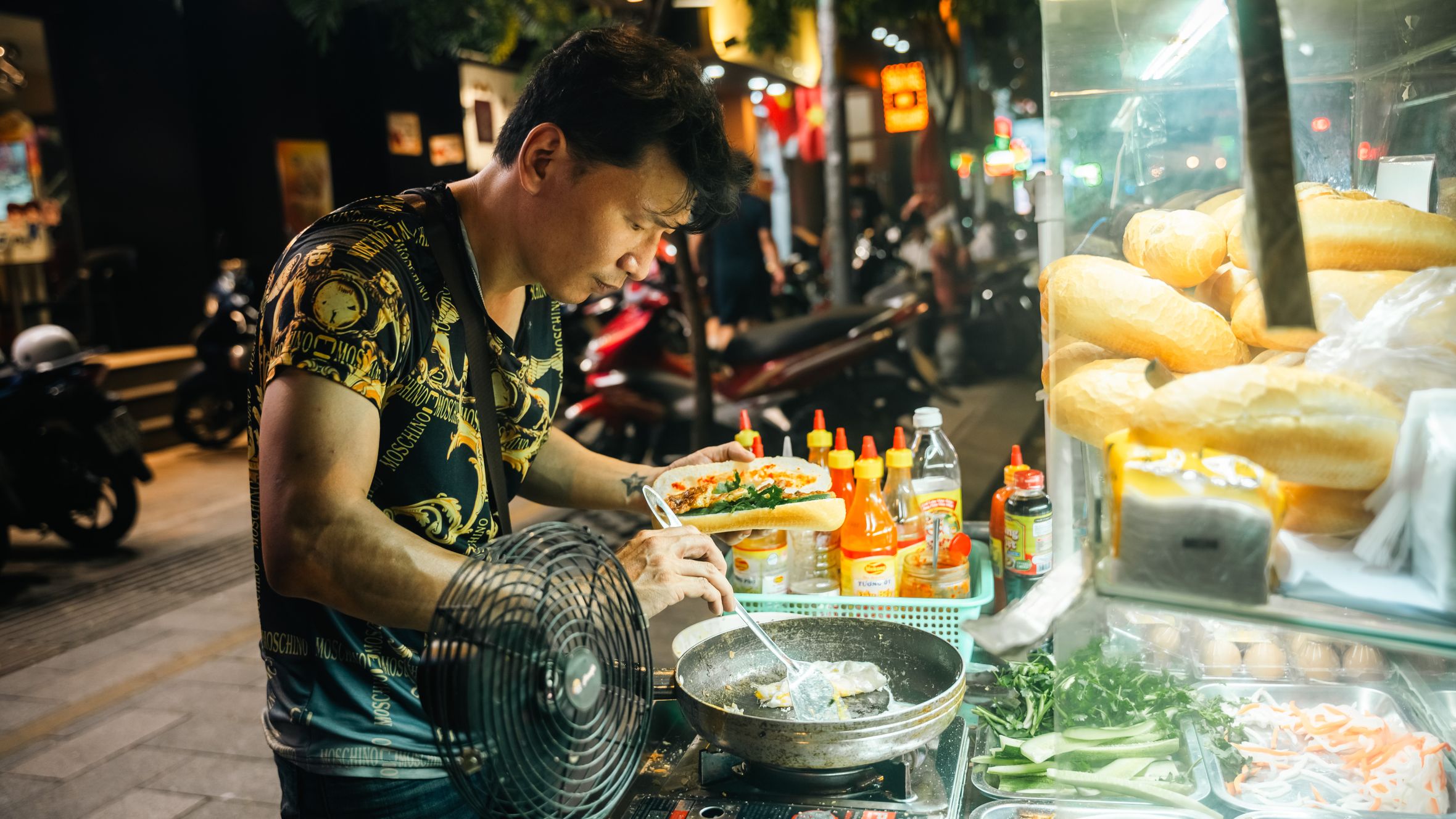 A street food vendor cooks and assembles Vietnamese banh mi at a bustling night market.