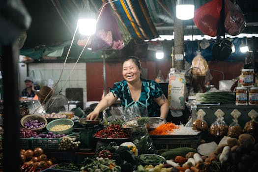 Smiling vendor at an Asian market stall displaying fresh vegetables and spices.