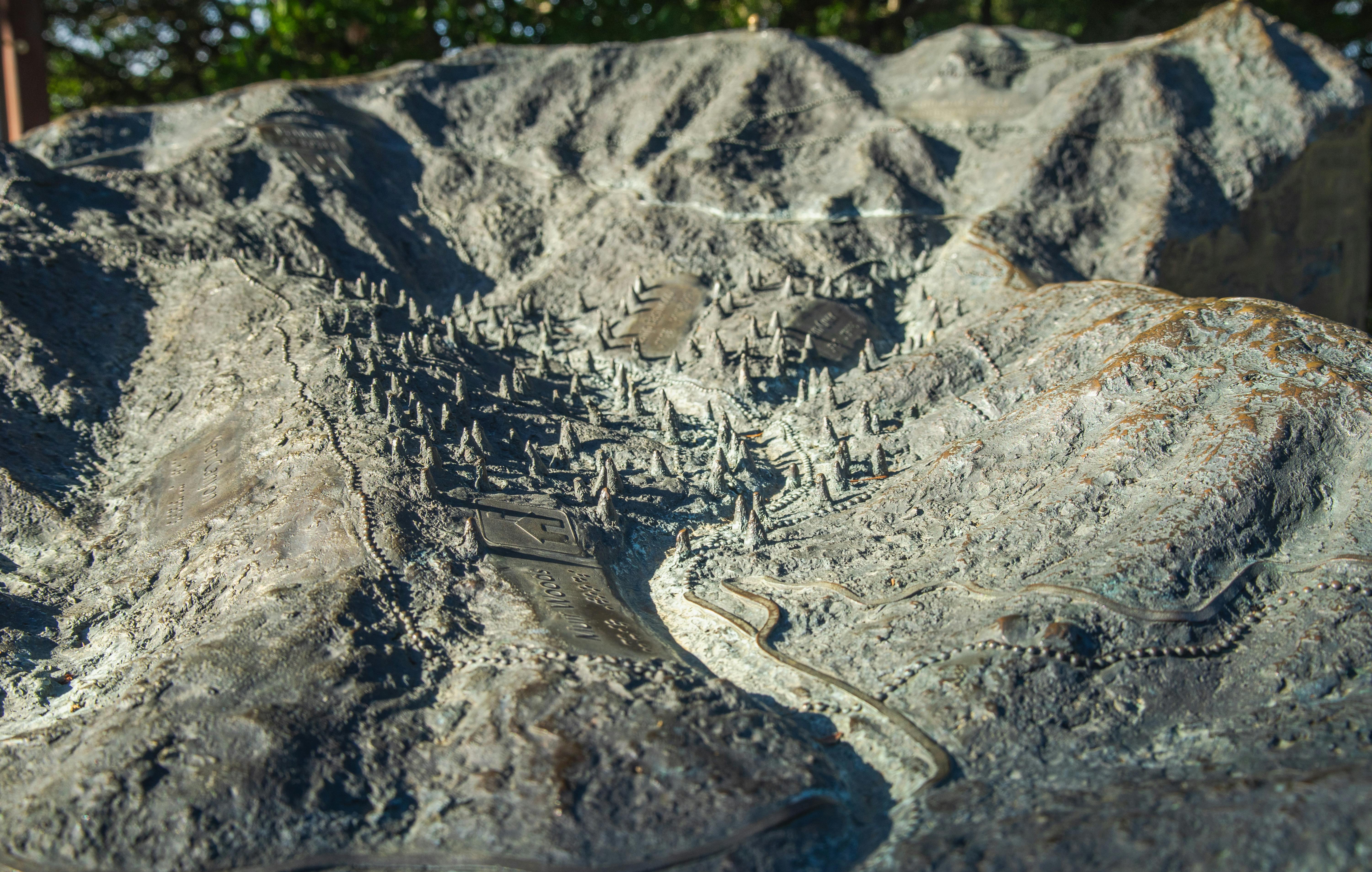 Free Detailed bronze relief map depicting mountainous terrain with a village and pathways under sunlight. Stock Photo