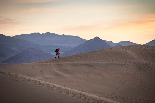 A lone hiker traverses sand dunes against a mountain backdrop at sunrise.