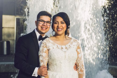 Free Smiling wedding couple in elegant attire posing by a fountain outdoors. Stock Photo