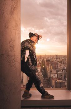 A stylish man poses against sunset skyline from a high vantage point indoors.
