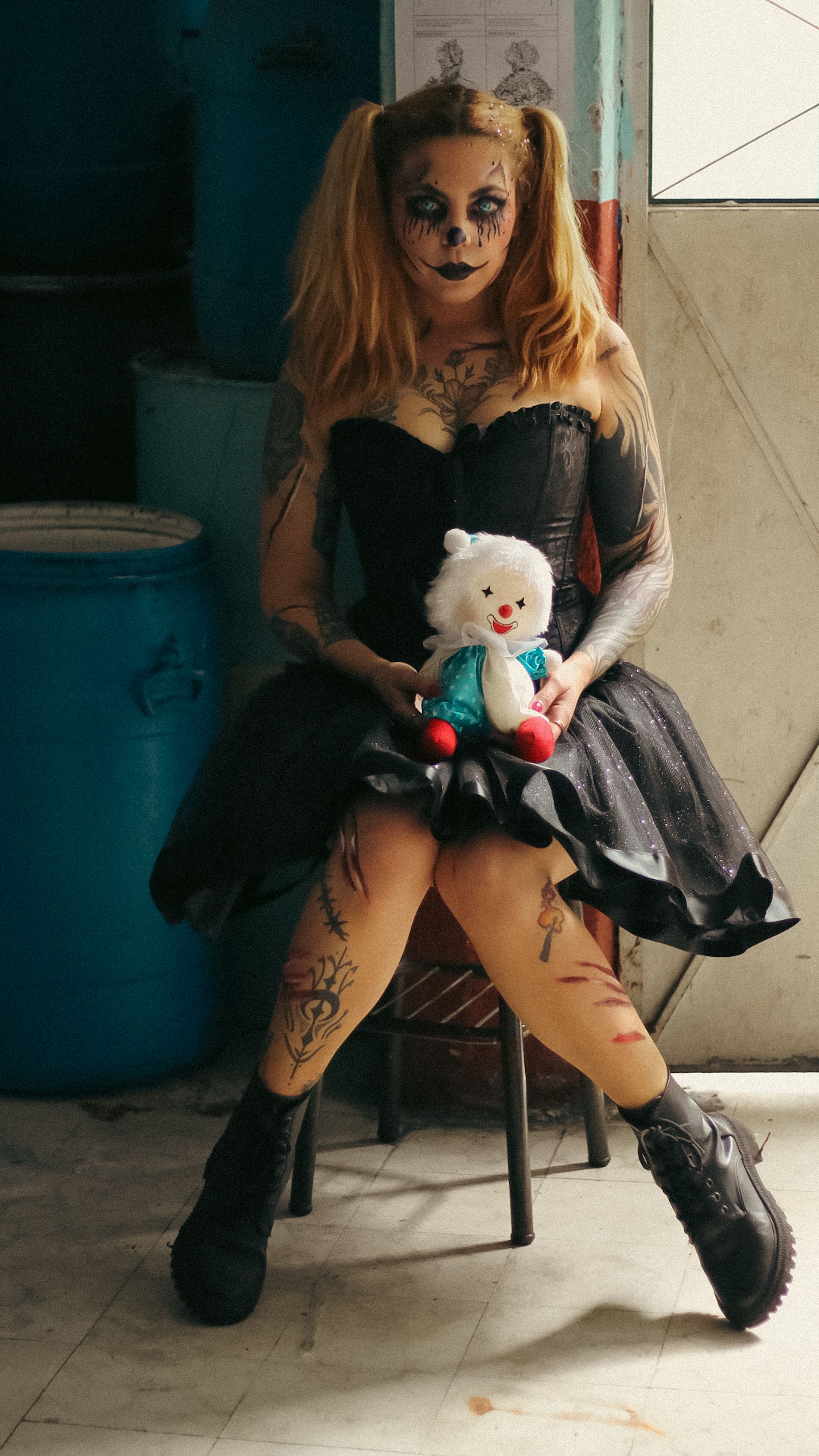 A person in gothic makeup and dress holding a toy bear, seated in an indoor setting.