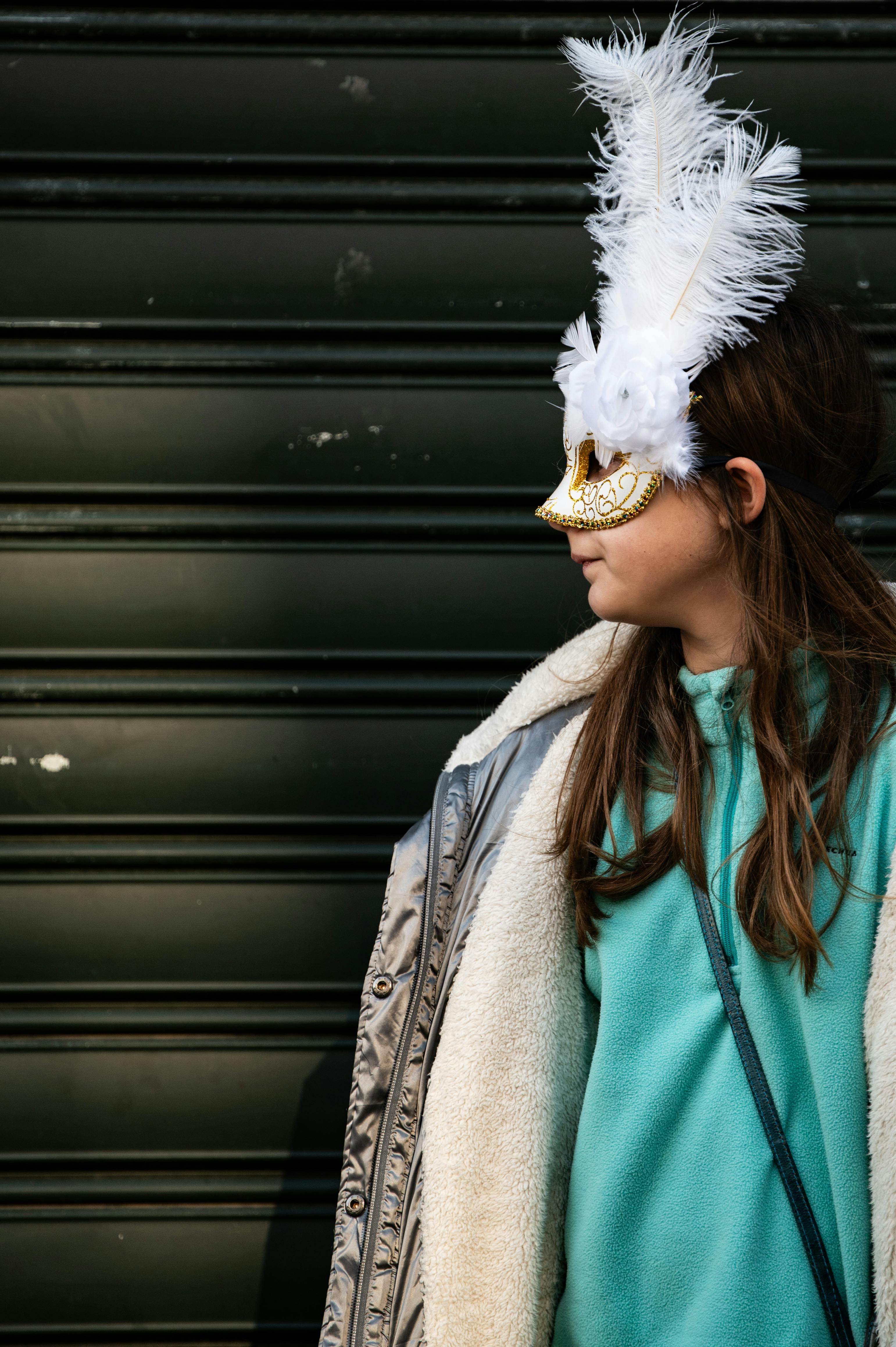 A girl wearing a feathered masquerade mask stands against a dark backdrop.