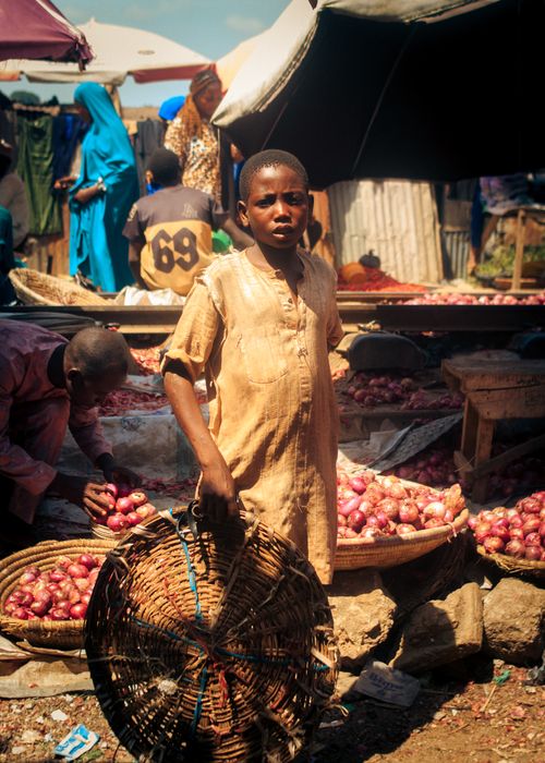 Free Young boy holding basket amidst a vibrant African market, with baskets of onions nearby. Stock Photo