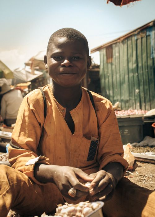 Free A cheerful young boy sits in an outdoor market, dressed in traditional attire, enjoying the lively environment. Stock Photo