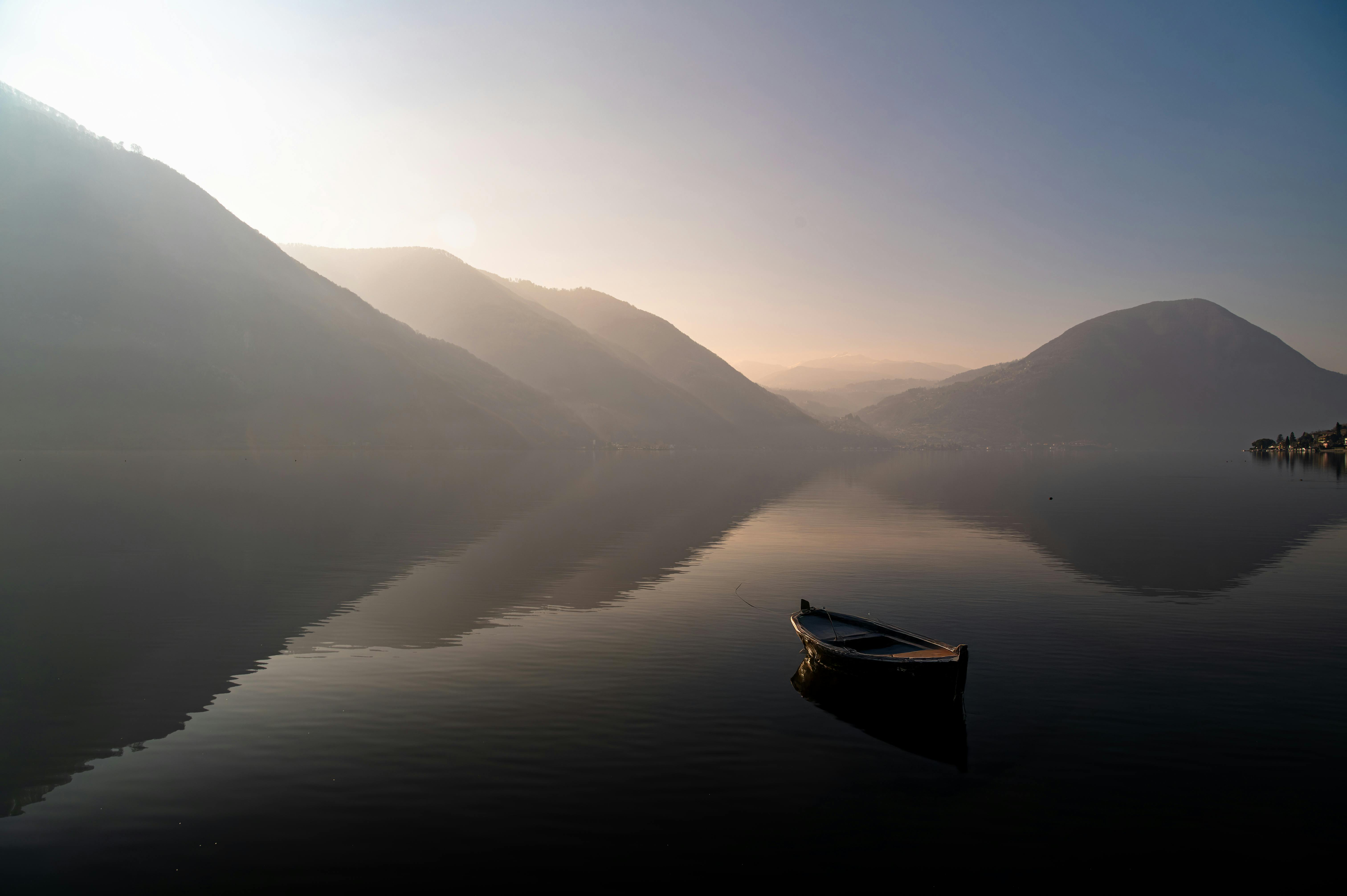 Calm water with a lone boat at sunrise, surrounded by misty mountains.