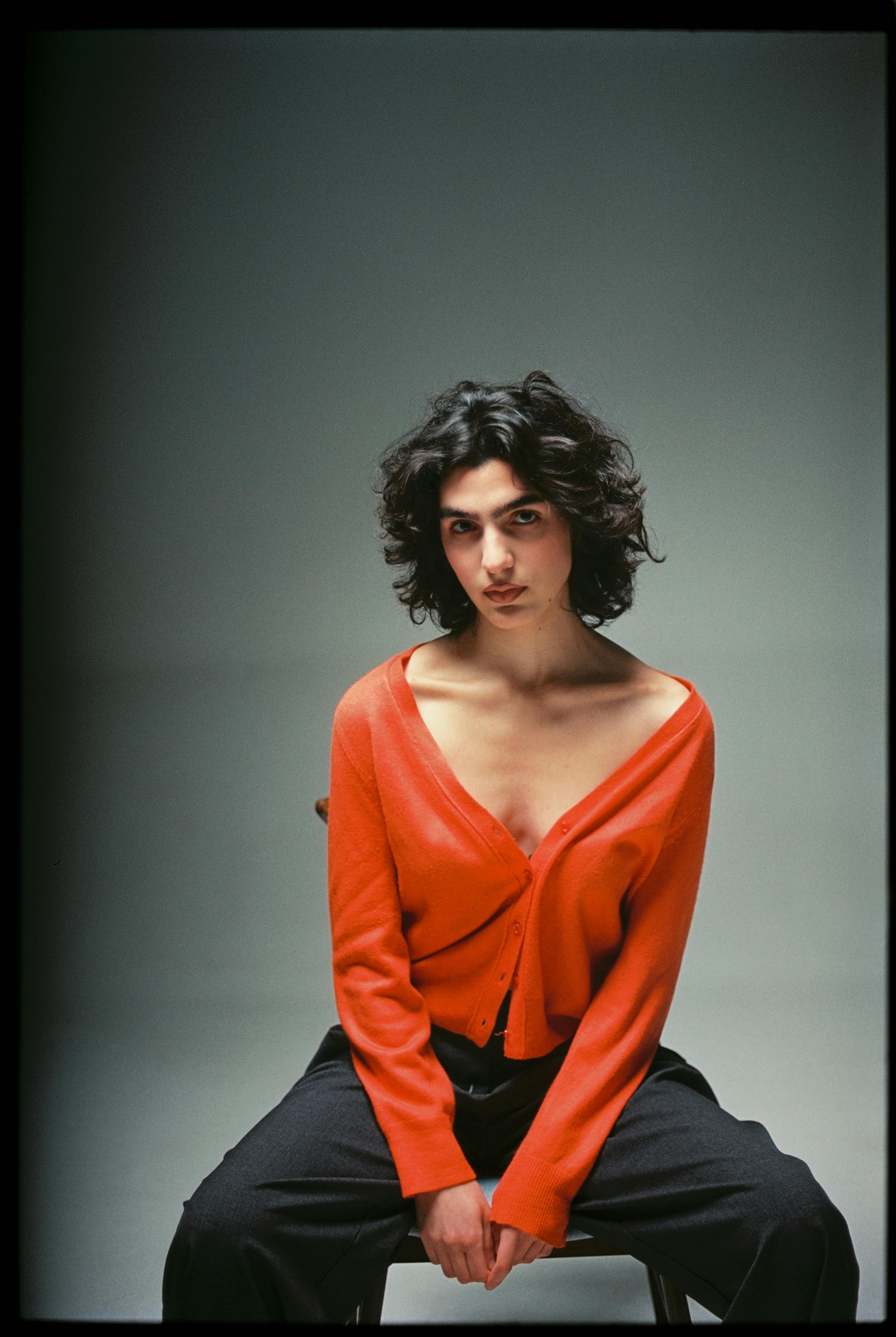 Fashion portrait of a confident woman in an orange cardigan sitting in a studio.