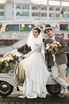 Charming bride and groom in wedding attire posing with floral-decorated scooter outdoors.