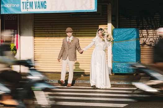 A couple in vintage wedding attire stands on a busy city street amidst moving scooters.