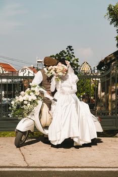 Bride and groom in vintage attire sitting on a classic scooter outdoors.
