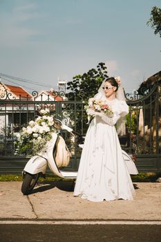 Stylish bride in sunglasses poses beside a decorated Vespa in a sunny outdoor setting.