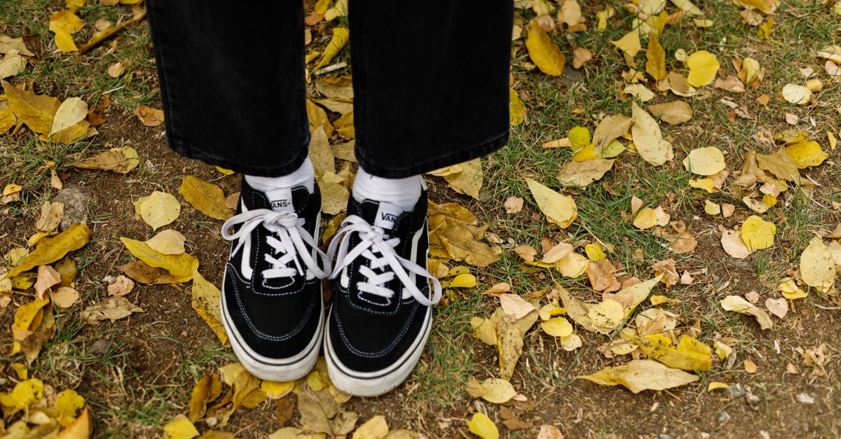 Low-angle shot of black sneakers with white laces on a grassy, leaf-covered path in autumn.