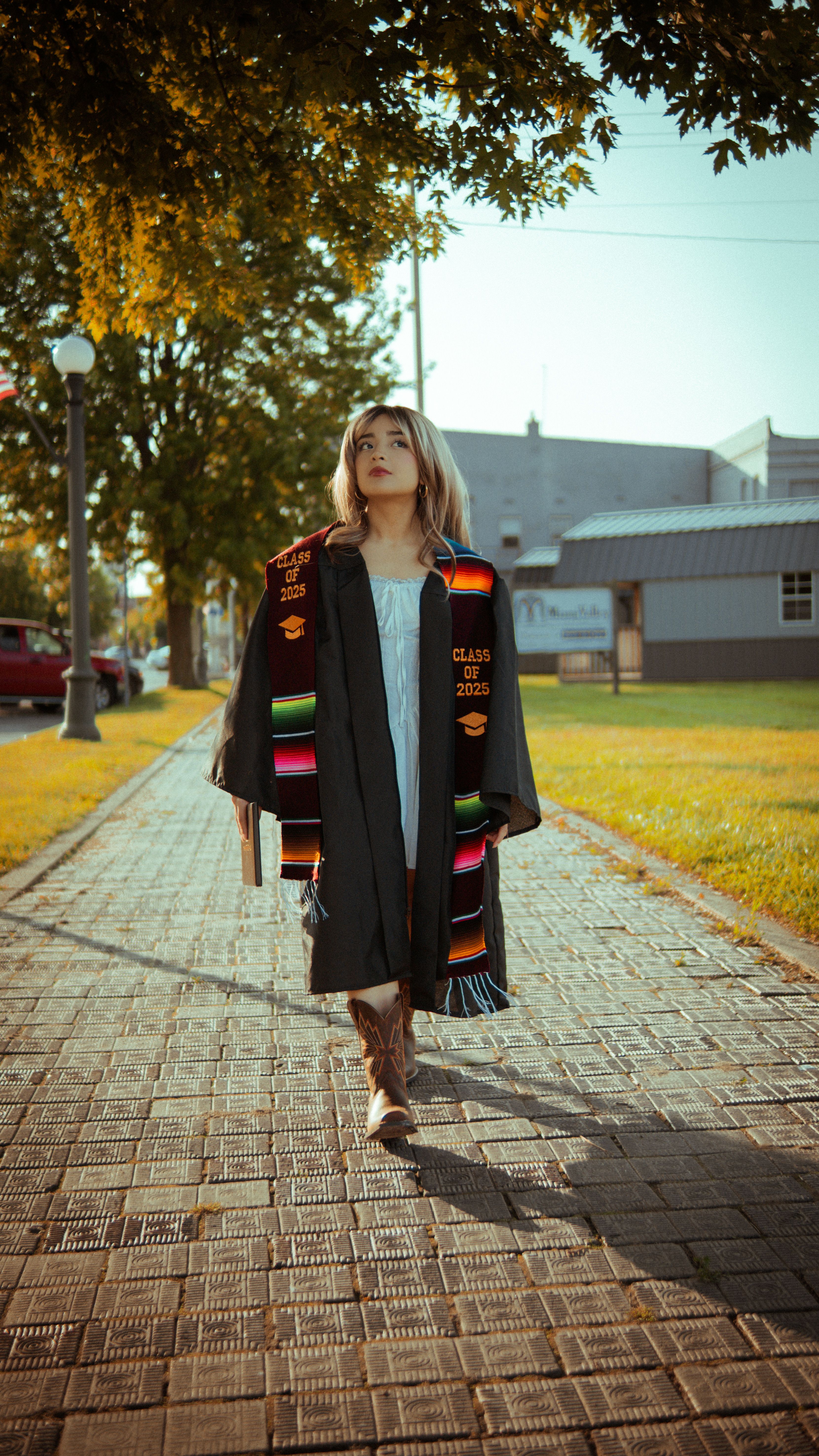 A young graduate walks confidently outdoors in a cap and gown with colorful sash, symbolizing accomplishment.
