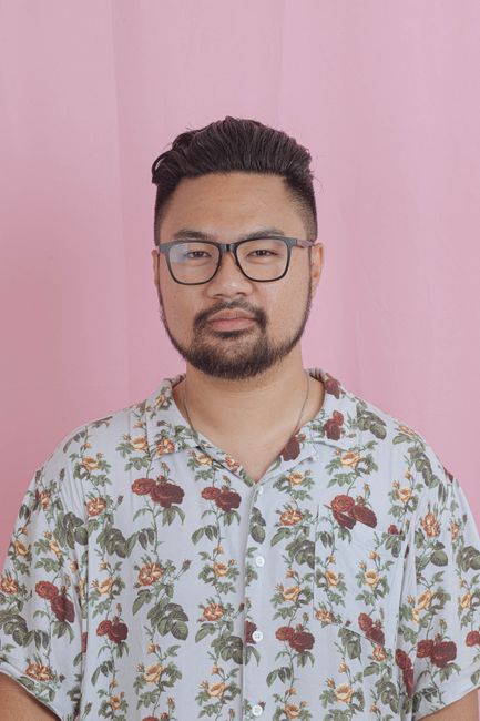 Close-up of a fashionable man wearing glasses and a floral shirt, set against a pink studio backdrop.