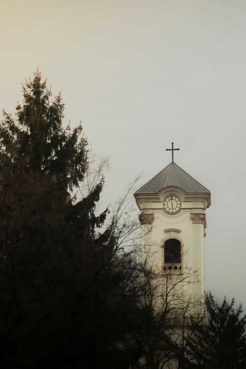 Free Classic church tower with cross against an overcast sky, next to tall trees. Stock Photo
