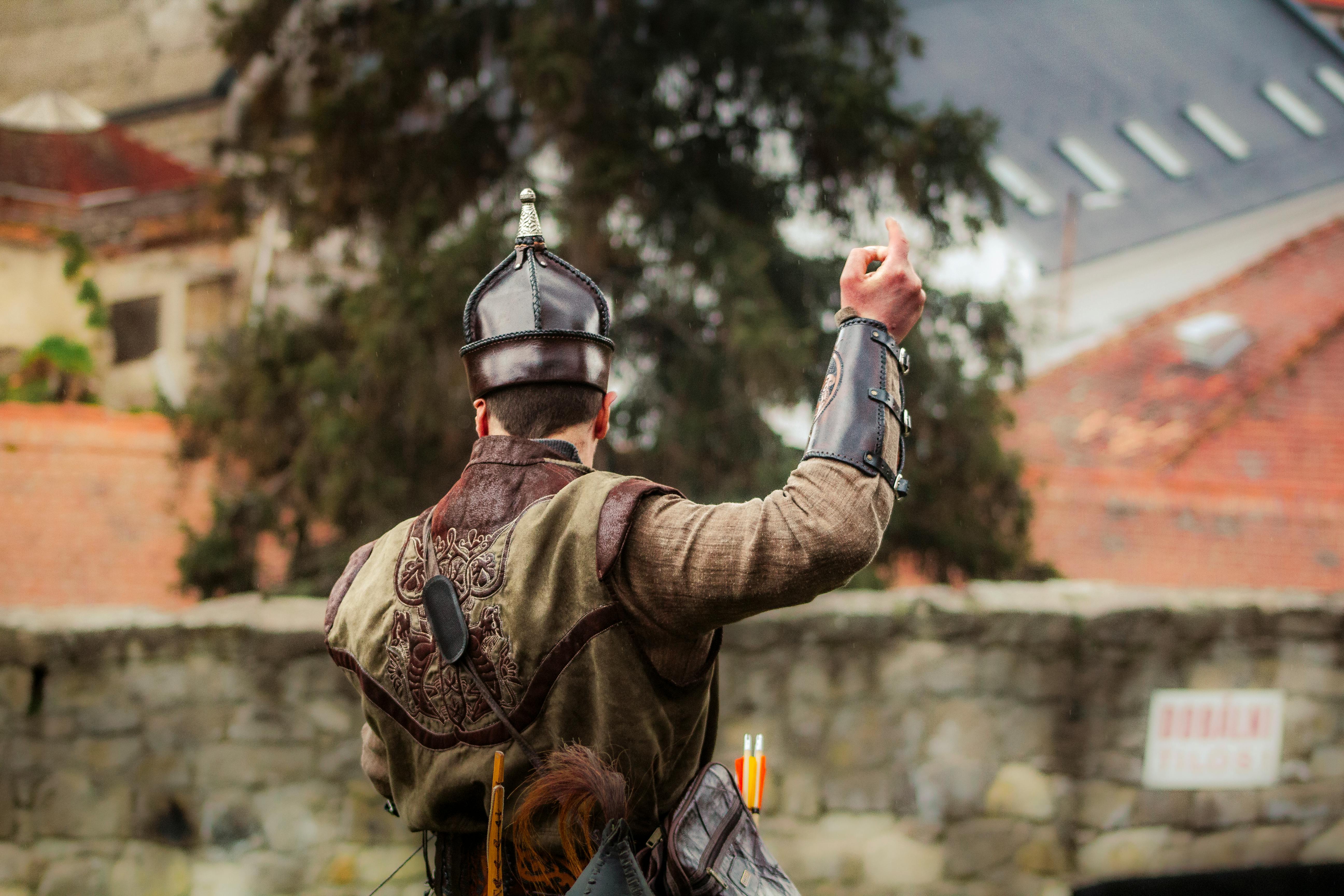 Medieval reenactor in armor with arrows and helmet facing a stone wall outdoors.