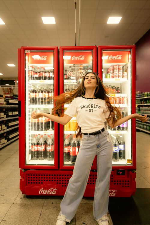 Free Confident woman in casual attire poses in front of a Coca-Cola fridge inside a store. Stock Photo
