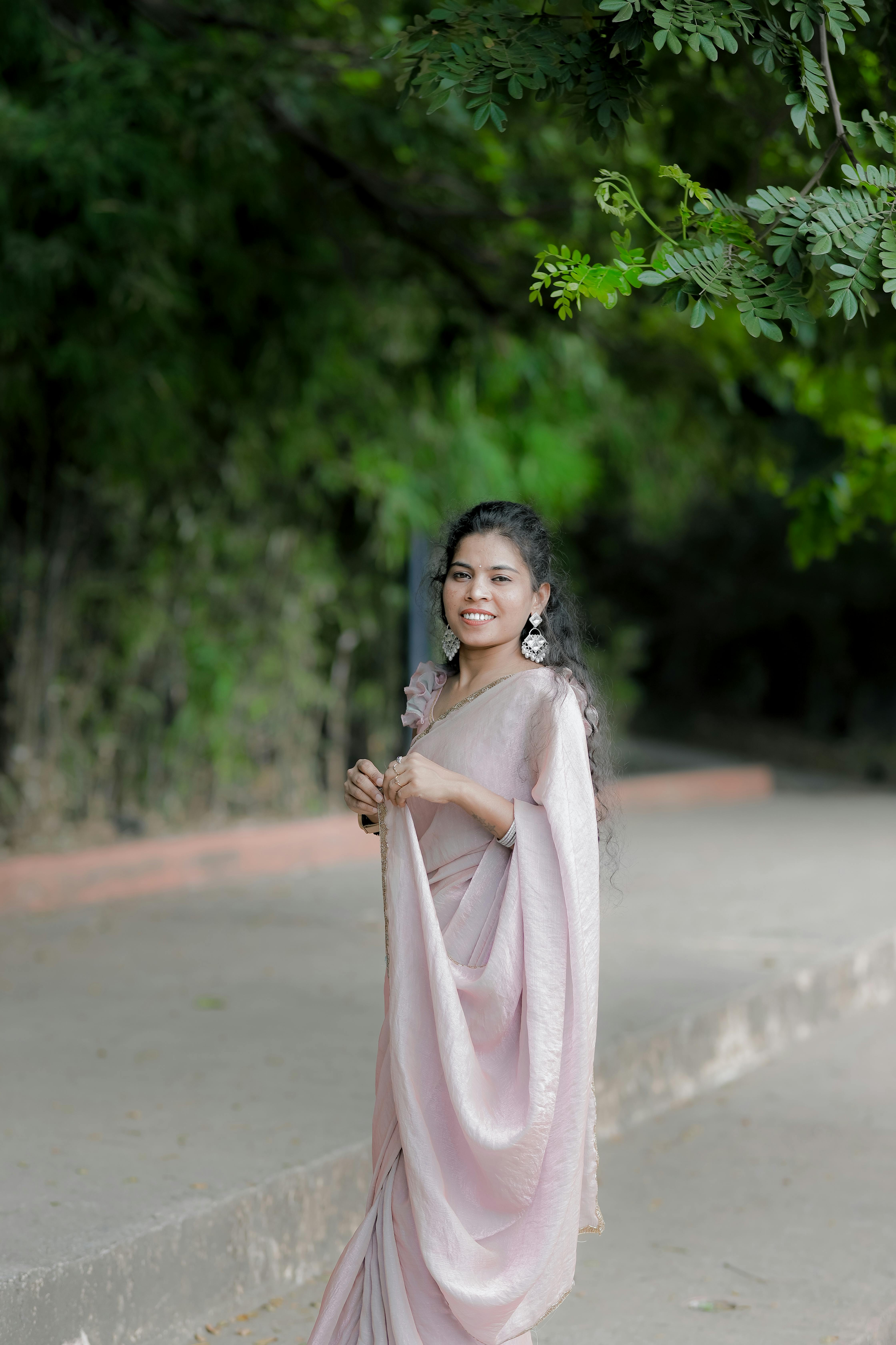 A woman in a pink saree, smiling and posing outdoors amidst lush greenery.