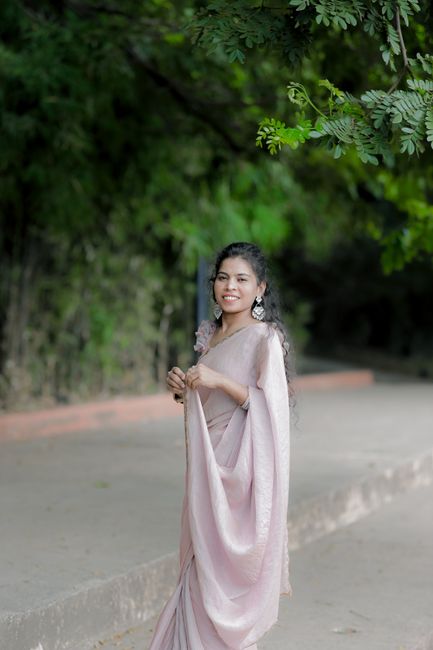 A woman in a pink saree, smiling and posing outdoors amidst lush greenery.