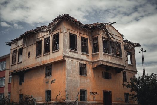 A dramatic view of an abandoned, decaying house with broken windows and damaged roof under a cloudy sky.