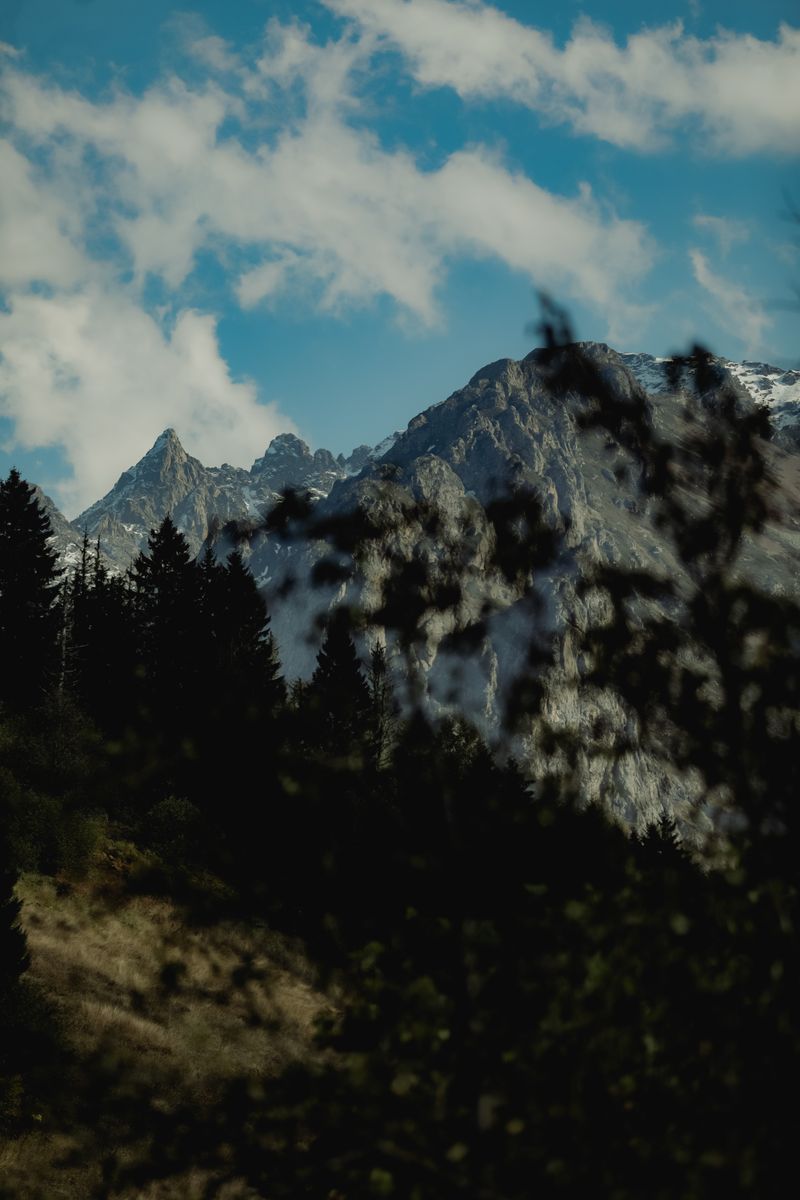 A captivating view of alpine mountains framed by lush trees and a vibrant cloudy sky.