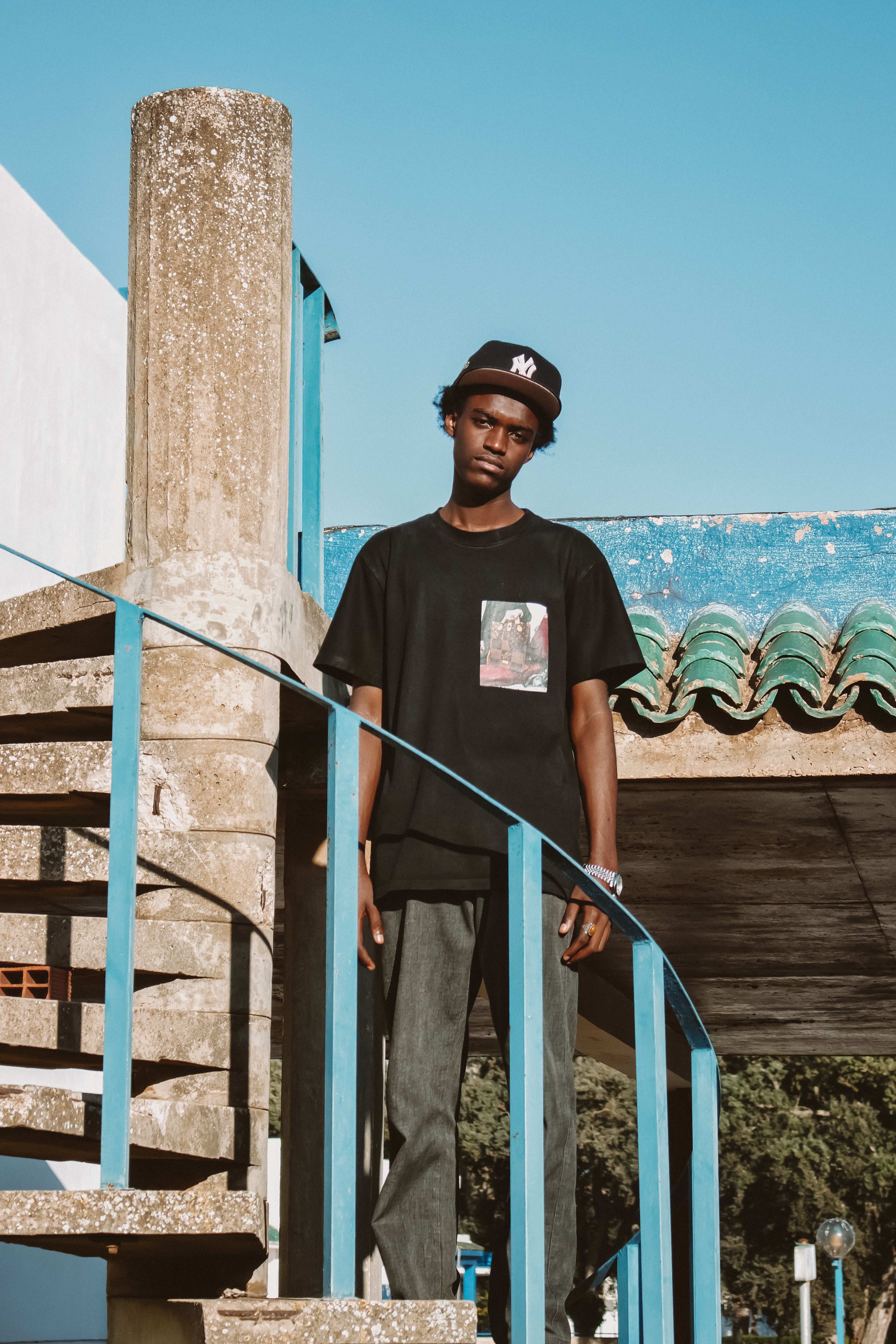 Retrato casual de um jovem encostado em uma grade em um ambiente urbano ao ar livre sob céus azuis claros.
