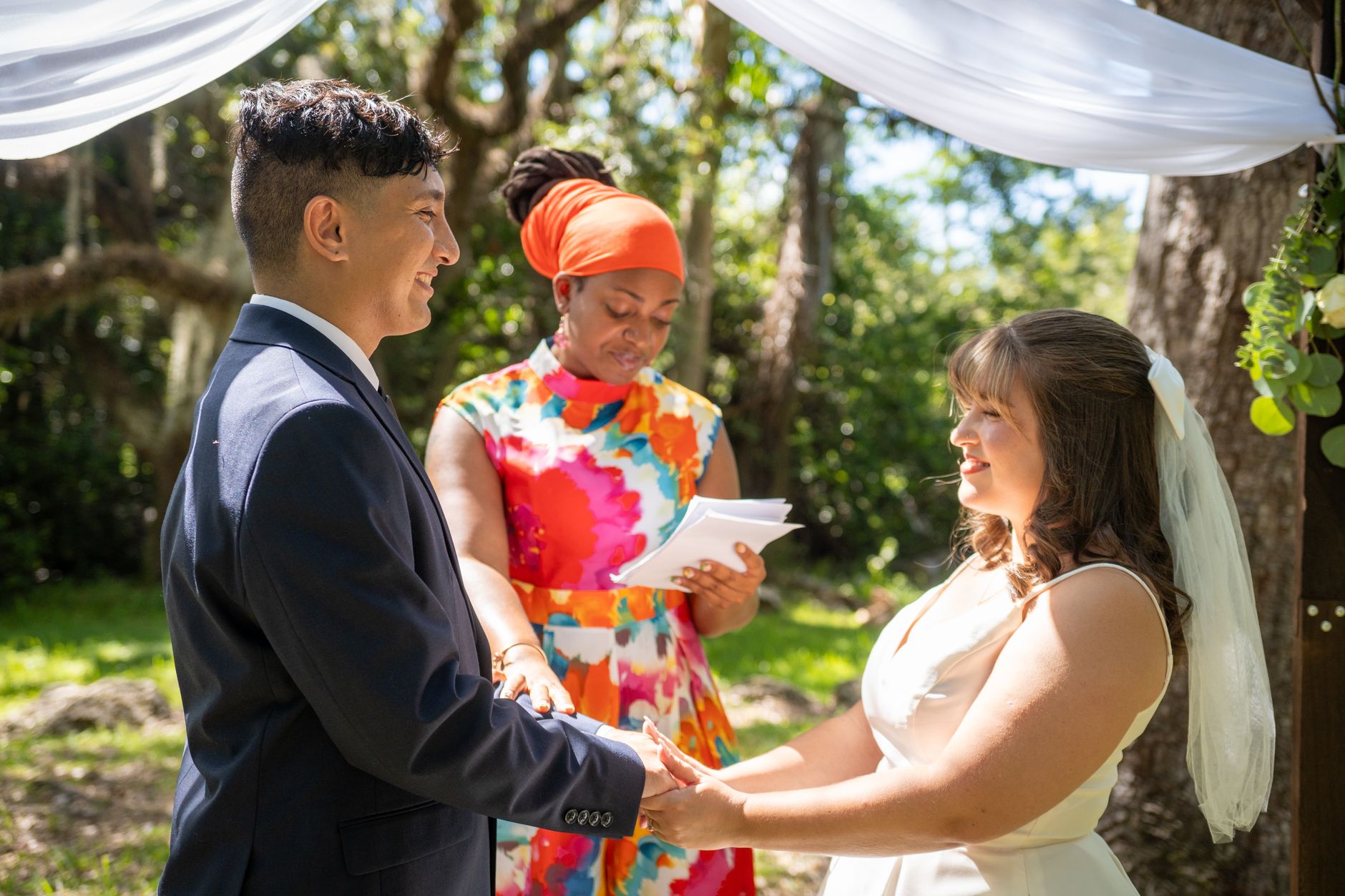 summer wedding trends egypt — Bright outdoor wedding under trees with officiant holding vows and couple exchanging rings.