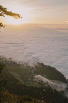 A stunning aerial view of a village submerged in dense fog during sunrise, framed by mountains and trees.