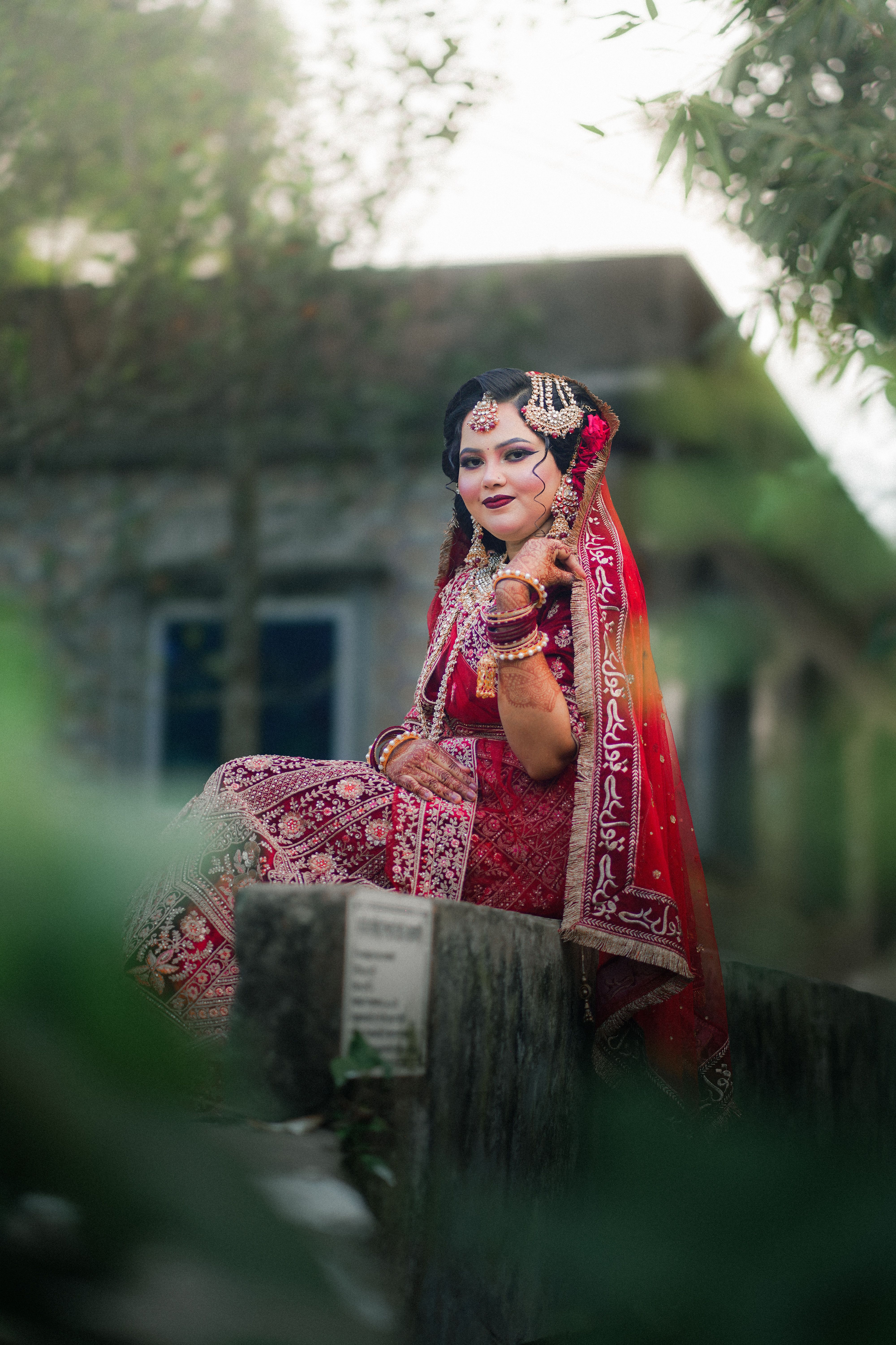 A traditional bride in vibrant red attire poses elegantly outdoors amidst natural surroundings.