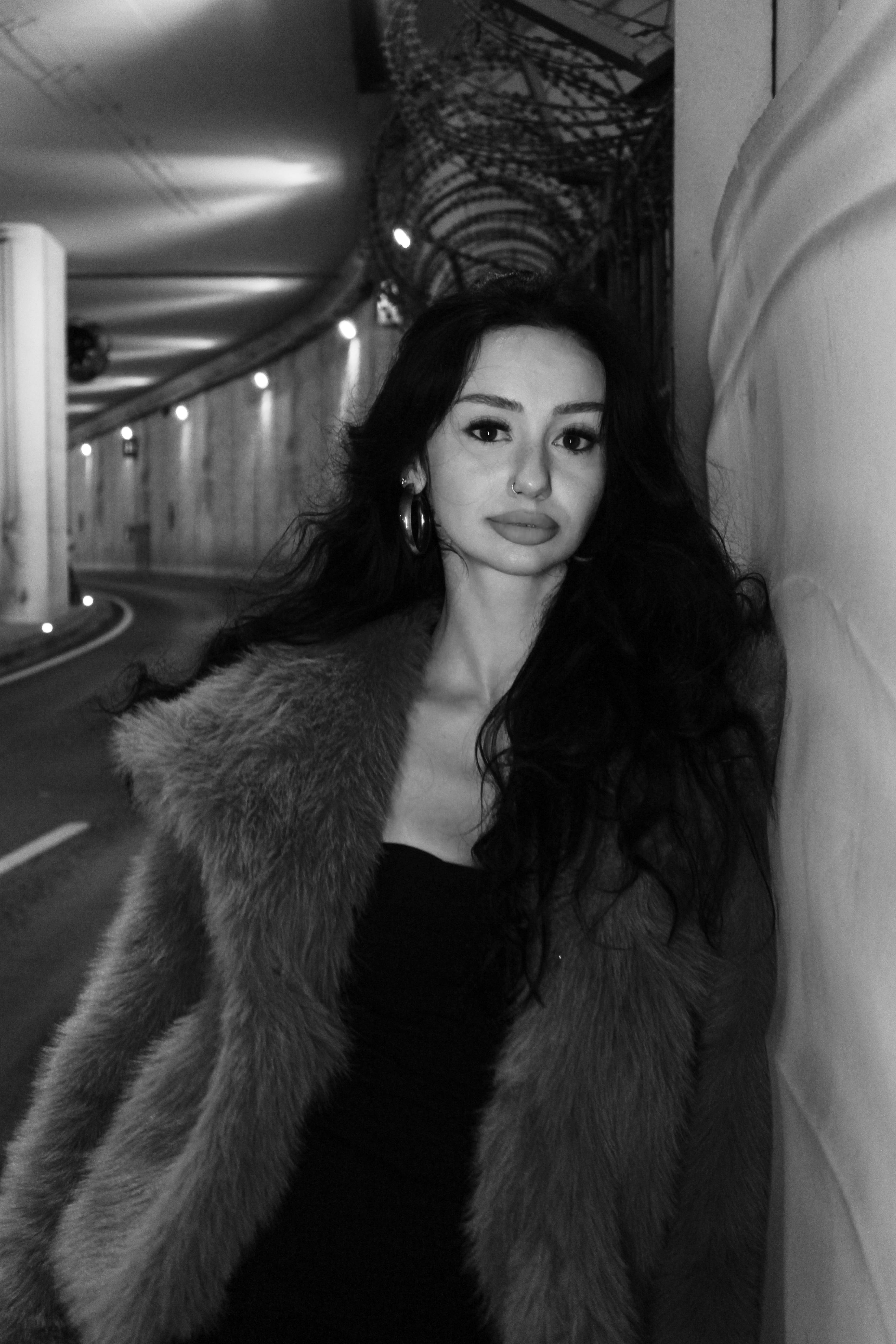 Elegant woman in a fur coat standing in an urban tunnel, captured in black and white.