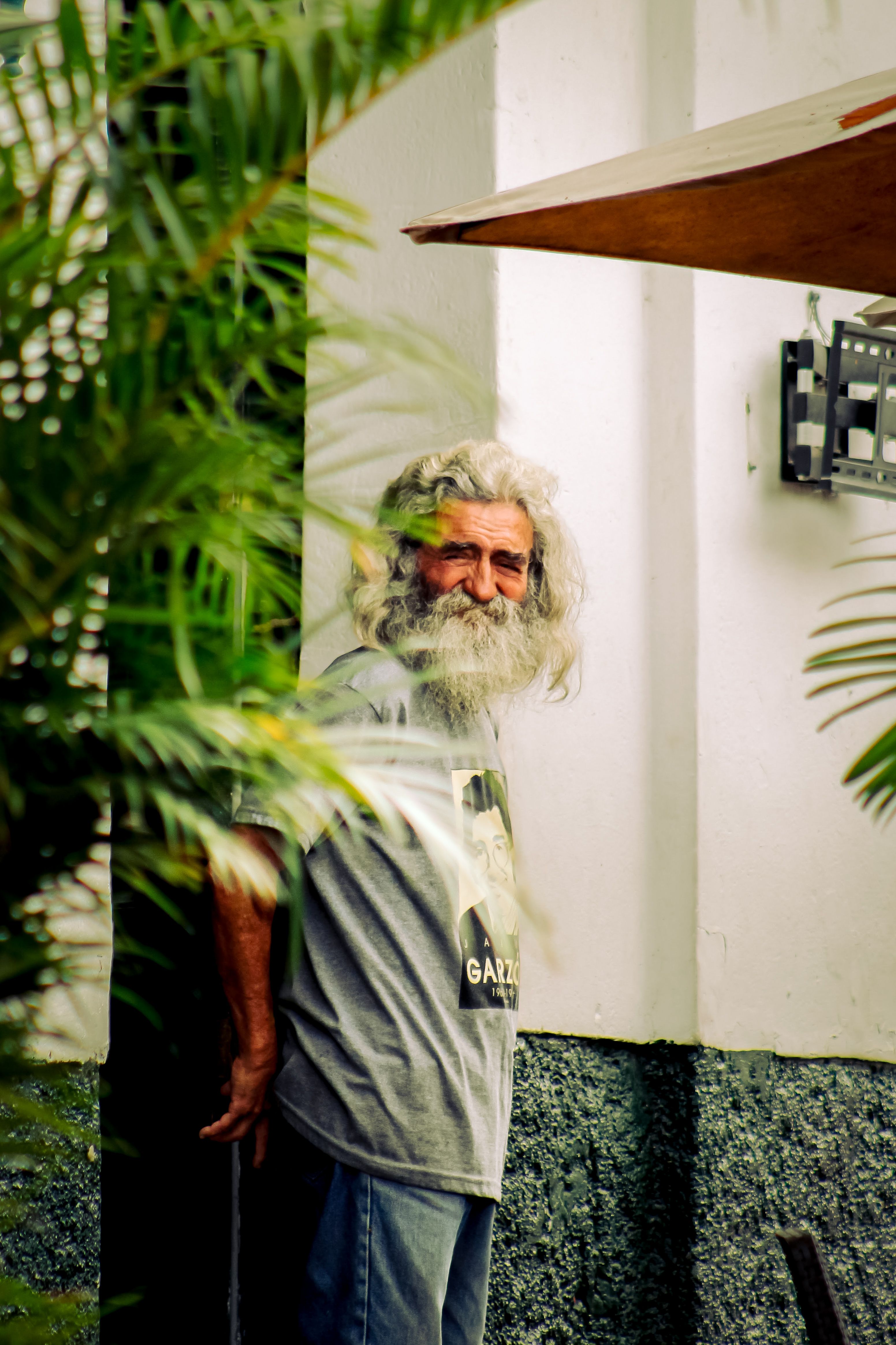 Elderly man with a long white beard standing near a wall, surrounded by green plants.
