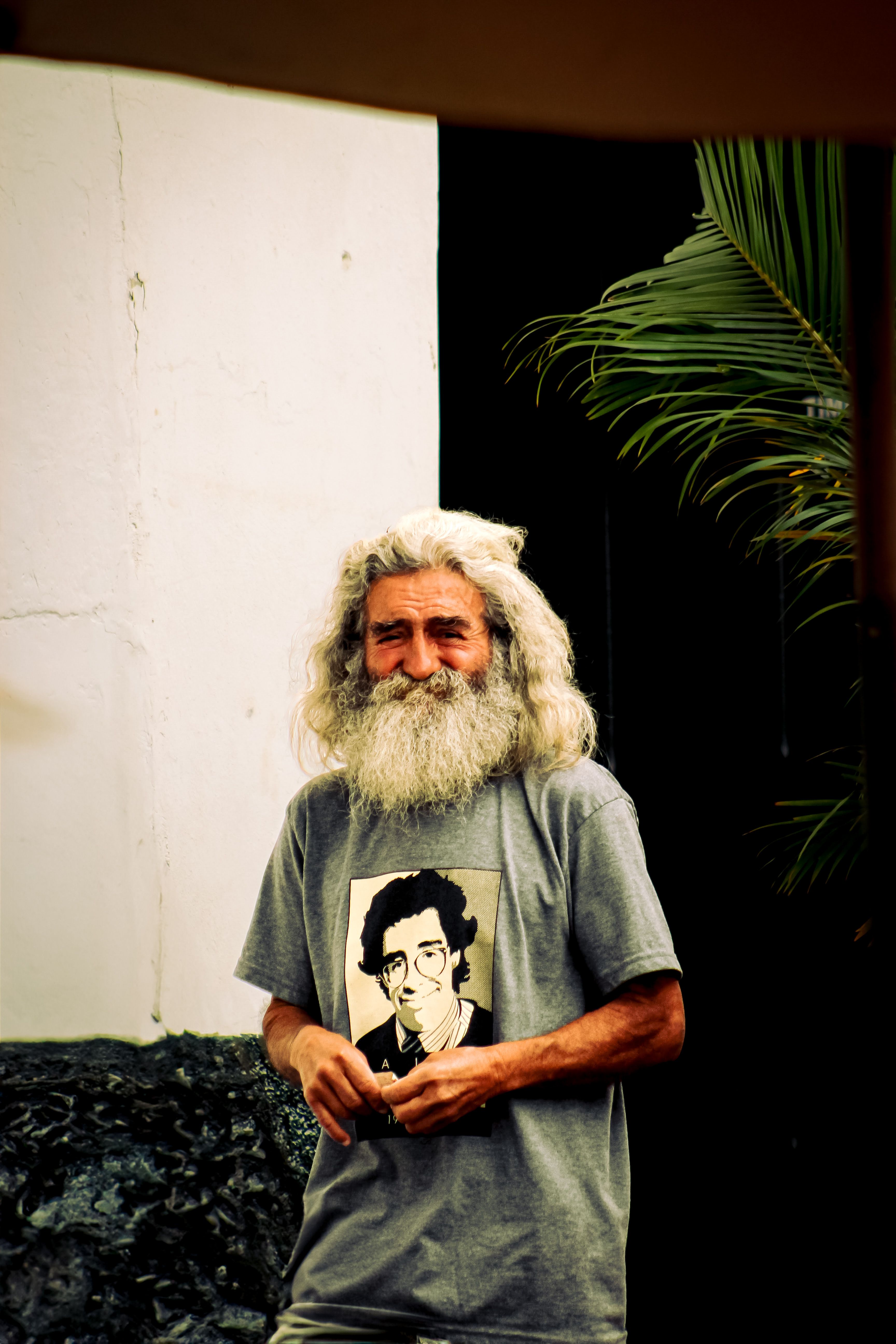 Elderly man with long beard wearing a t-shirt featuring a portrait, standing outside near a white wall and palm tree.
