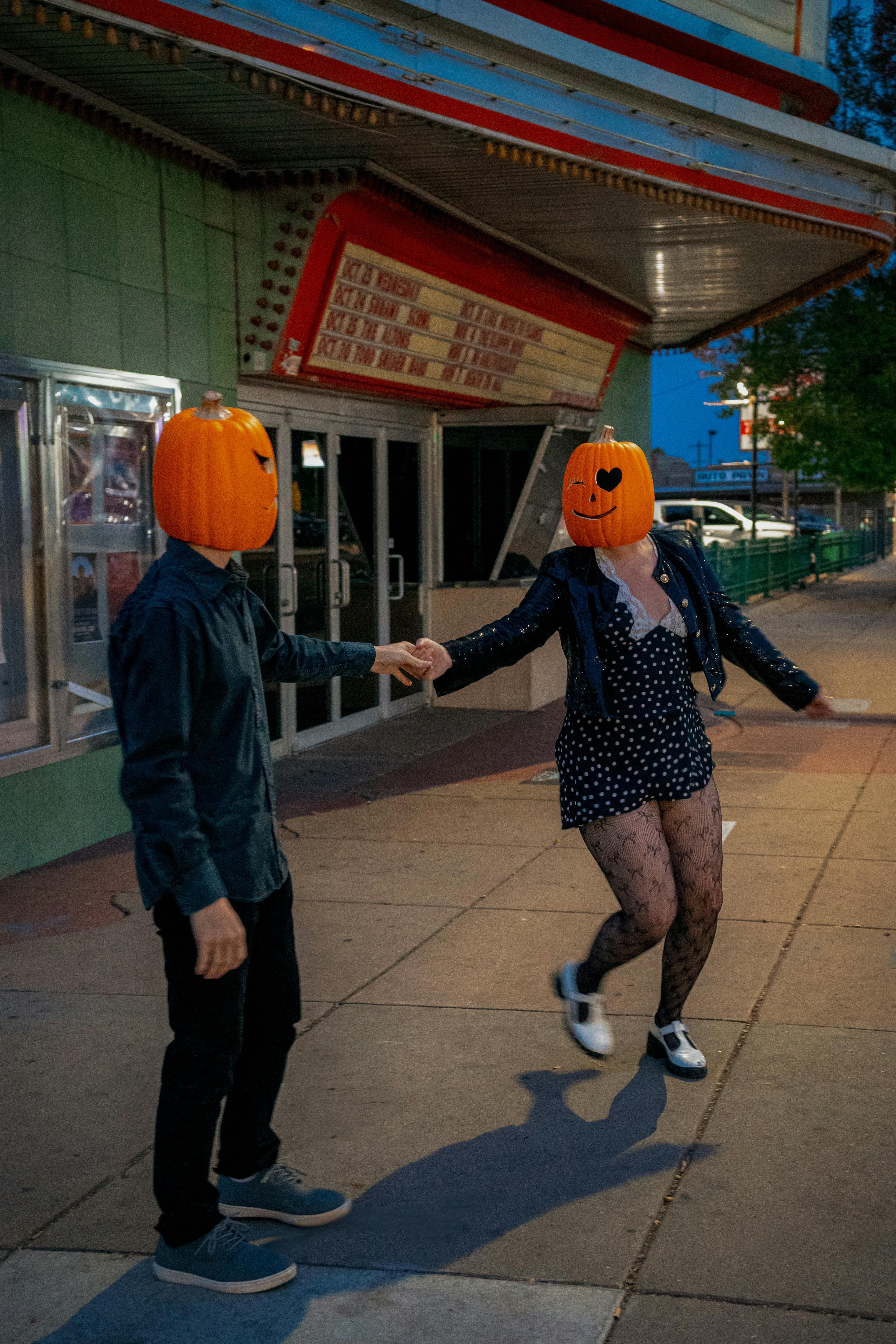 Free A playful couple with carved pumpkin heads dancing outside a vintage theater in fall. Stock Photo
