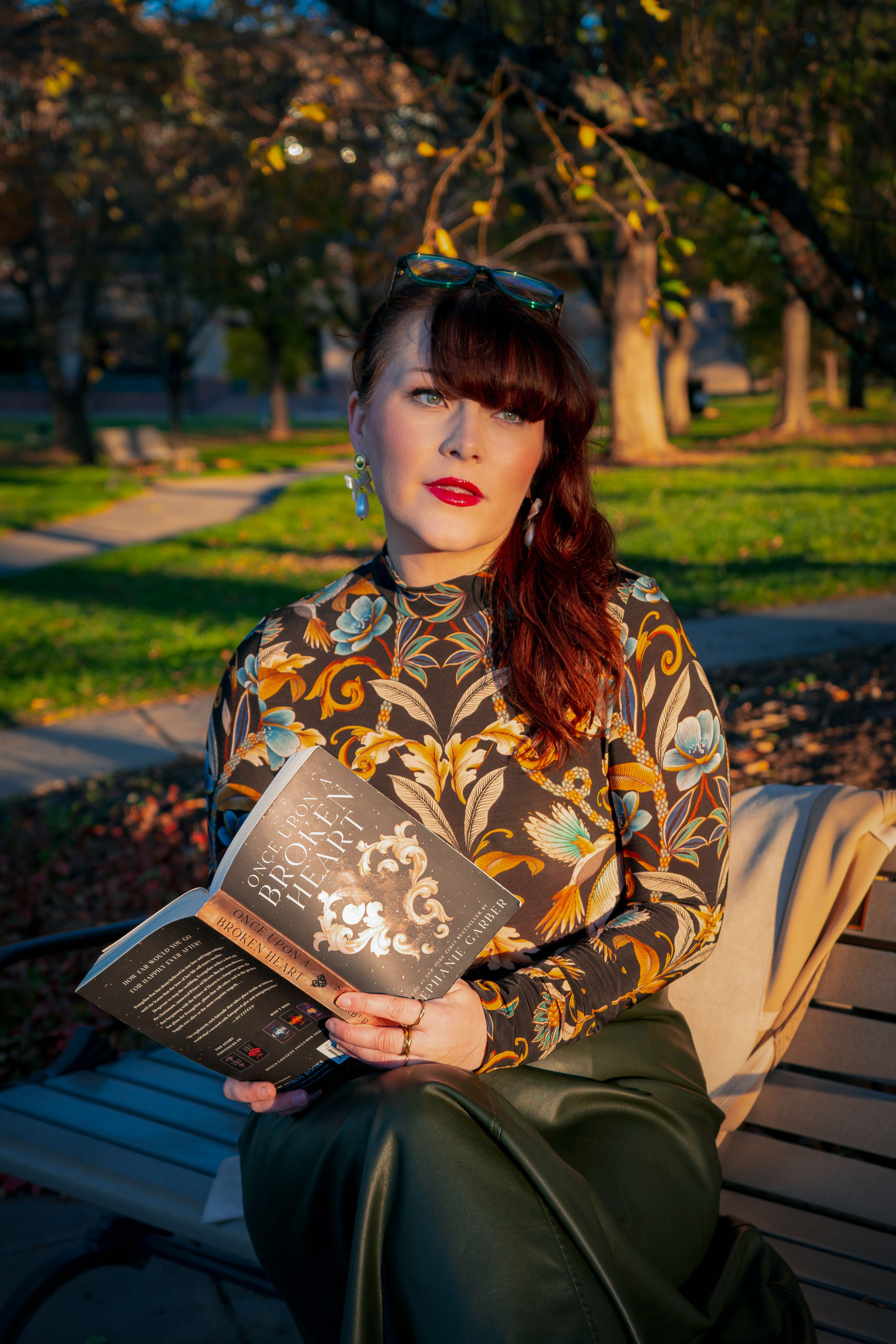 Stylish woman reading in a park during fall, highlighted by vibrant outfit and lush foliage.